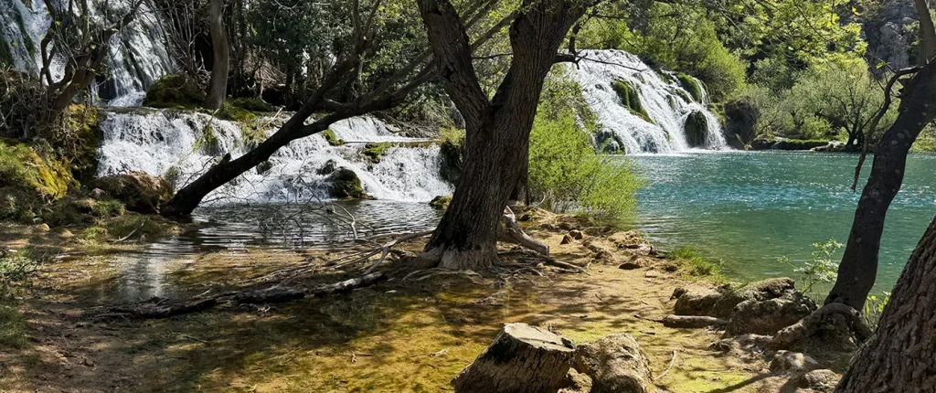 Sunlit waterfalls cascading into turquoise waters of the Krupa River, surrounded by lush trees, moss-covered rocks, and shallow streams.